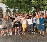 Fröhliche Gruppe mit jungen Menschen posiert im Freien, Arme oben. Im Hintergrund ein Gebäude.