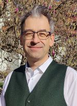 Person im Porträt, fotografiert vor blühenden Ästen, trägt formelle Kleidung.