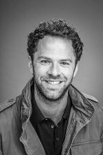 Porträt einer lächelnden Person mit dunklen Locken und Bart, gekleidet in eine Jacke.