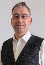 Person im Porträt, fotografiert vor blühenden Ästen, trägt formelle Kleidung.