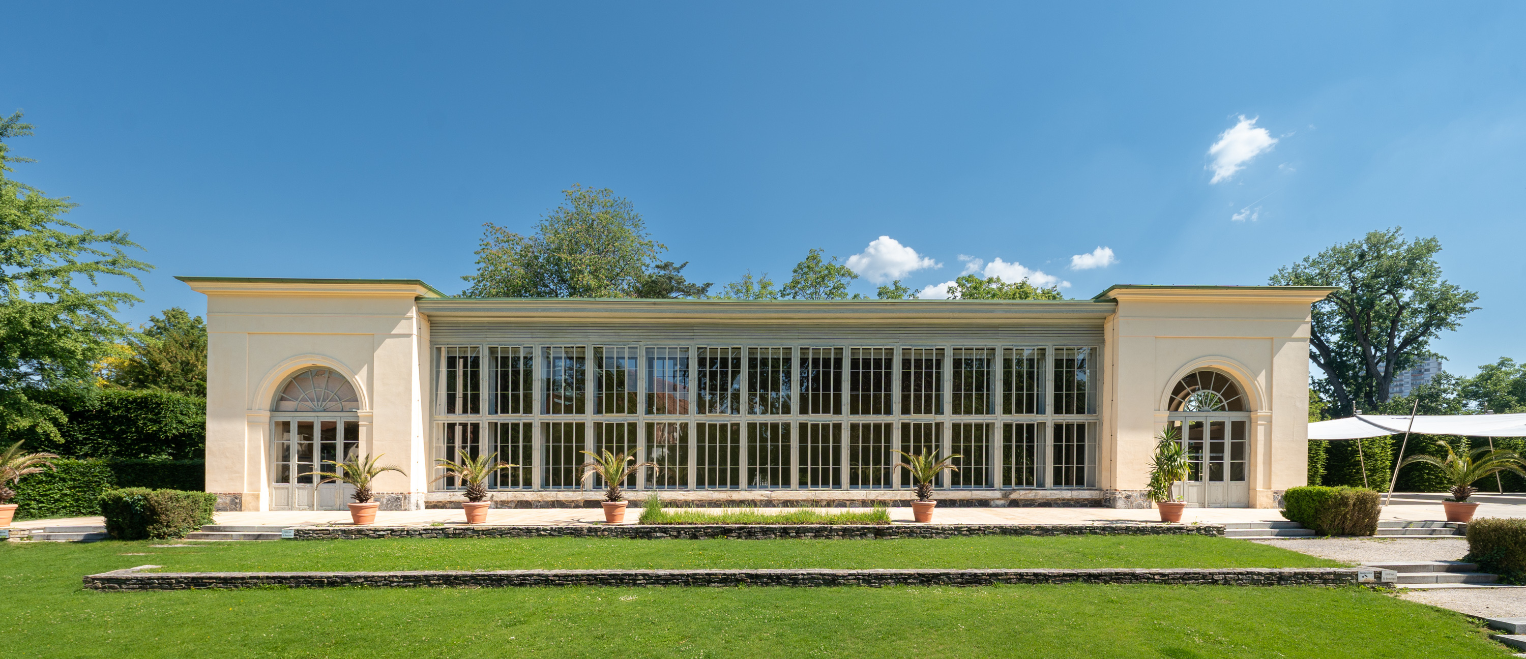 Historischer Pavillon mit grosser Fensterfront, umgeben von Grünflächen und Pflanzen.