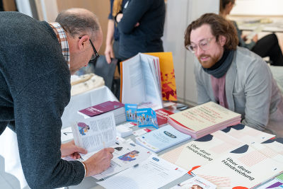 Impressionen von den Marktständen, ein Mann notiert sich Informationen, vor ihm eine Menge an Broschüren