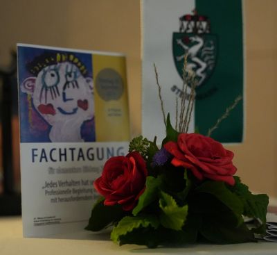 Ein Bild der Fachtagung. Es zeigt einen Land Steiermark Wimpel, die Fachtagungs-Einladung sowie ein Blumengesteck.